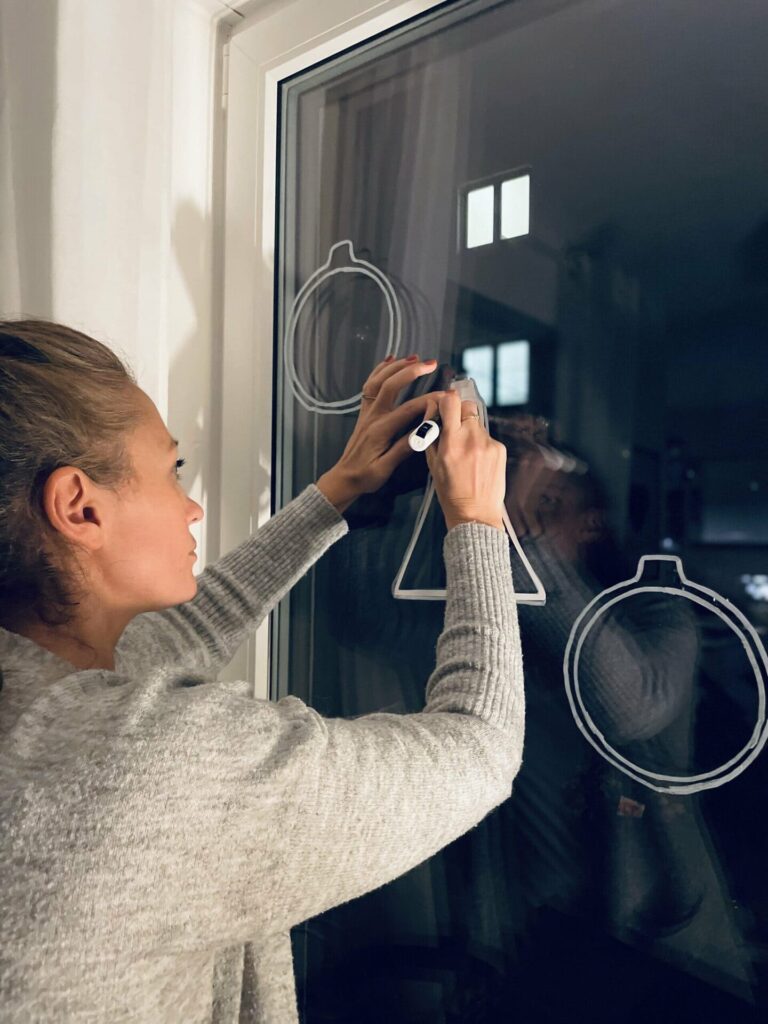 Eine Frau malt mit einem weißen Stift Symbole auf eine Fensterscheibe. Hinter der Scheibe ist es dunkel, da das Bild abends aufgenommen wurde.