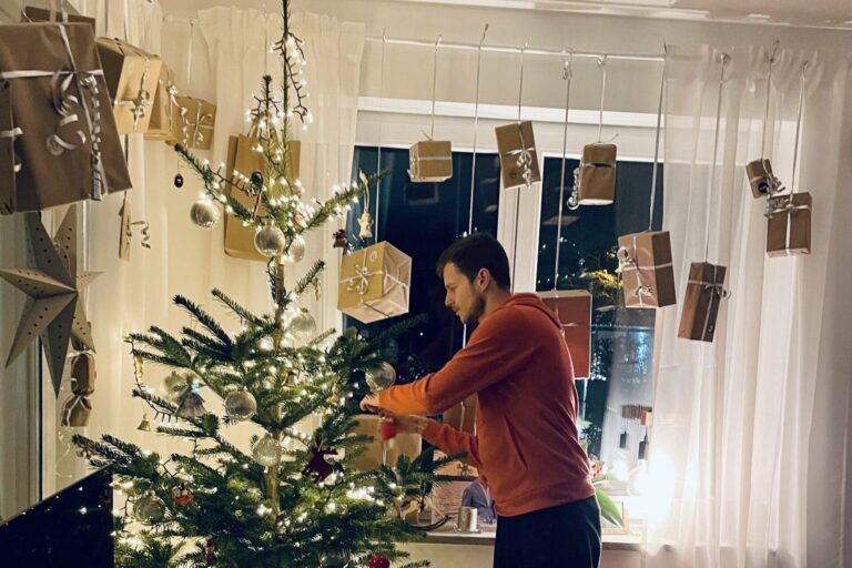 Ein Mann schmückt einen Weihnachtsbaum im Haus. Im Hintergrund hängen Weihnachtsgeschenke an vielen Schnüren an der Vorhangstange des Fensters.