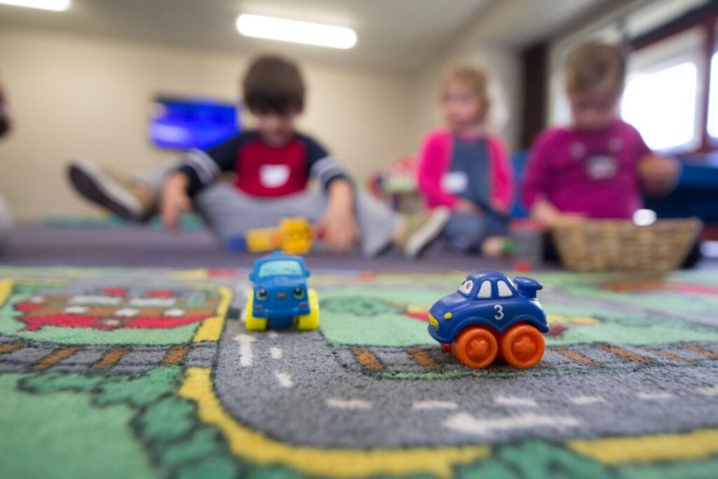 Zwei Spielzeugautos auf einem Spielteppich. Im Hintergrund sind drei Kinder zu sehen