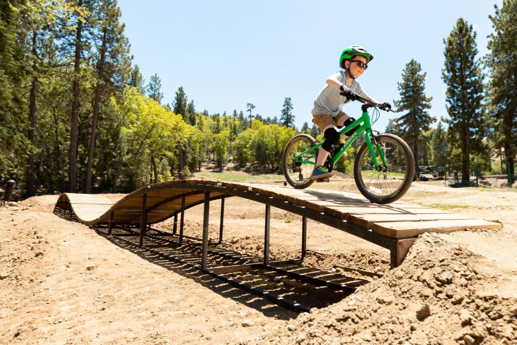 Sand und eine Brücke, im Hinterbrund Bäume. Auf der Brücke fährt ein Kind mit einem Fahrrad