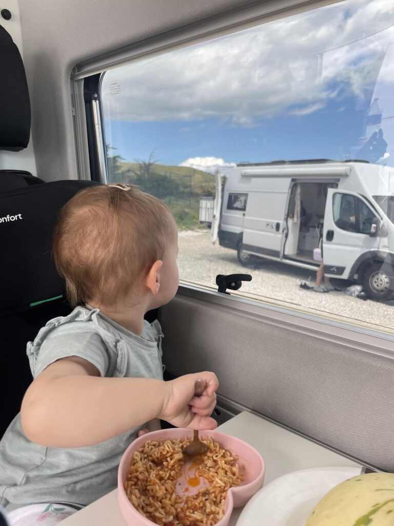 Kind sitzt im Auto am Tisch und isst Nudeln aus einem Kinderteller. Dabei sieht es seitlich zum Fenster hinaus. Im Hintergrund ein Wohnmobil