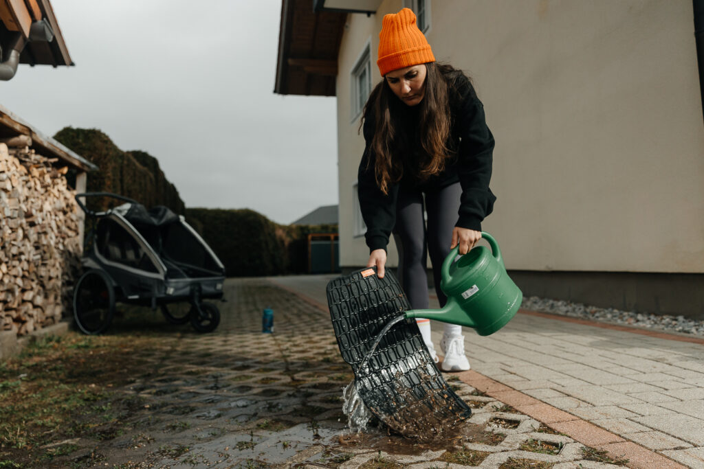 Eine Frau auf dem Parkplatz vor dem Haus säubert eine Schmutzfangmatte für den Fahrradanhänger indem sie Wasser aus einer grüßen Gießkanne darüber gießt